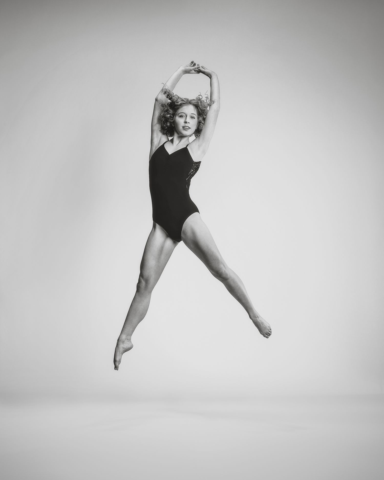Black and white photo of a female dancer leaping gracefully in a black leotard against a white background.