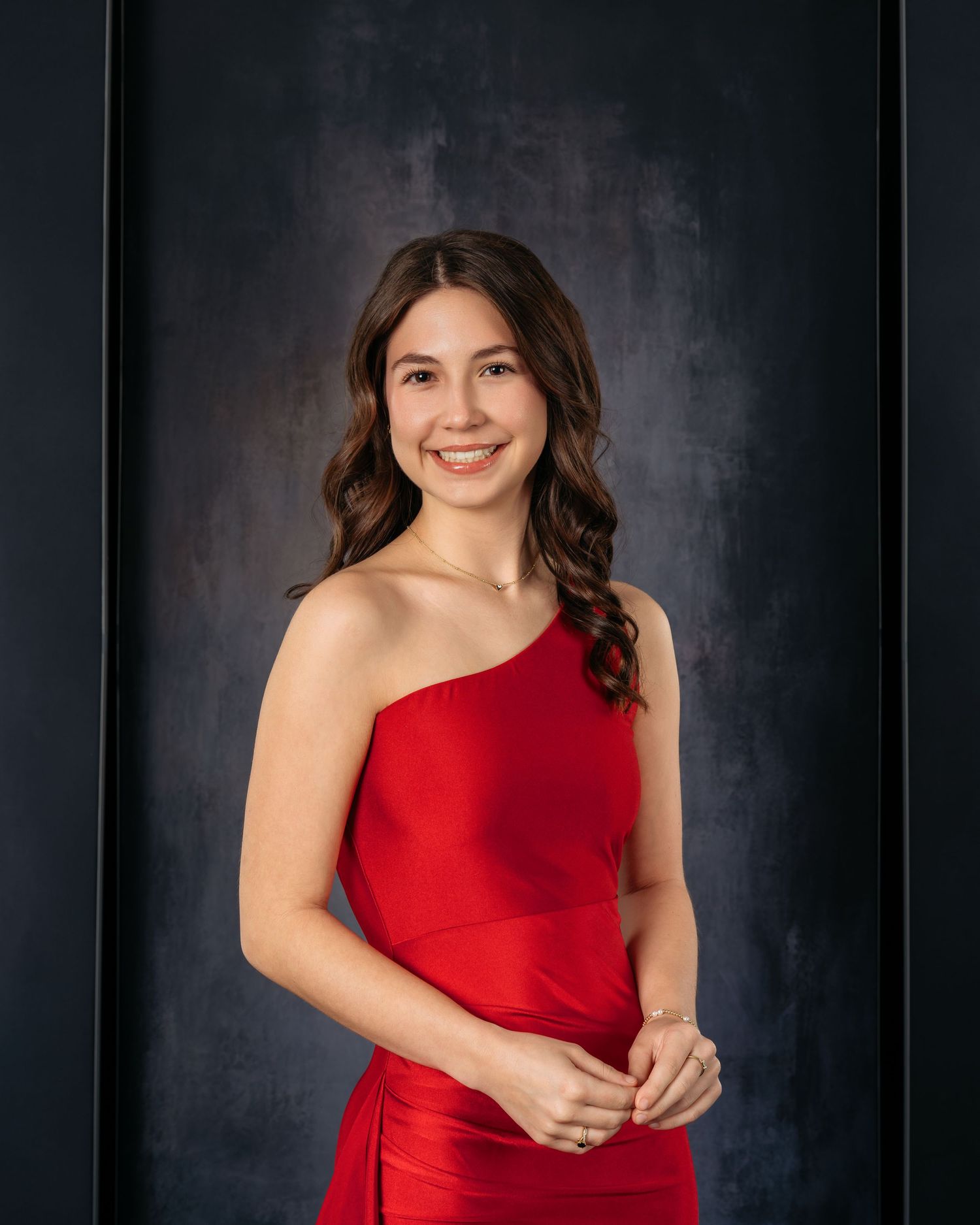 Young woman in elegant red one-shoulder gown smiling gracefully against dark studio backdrop.