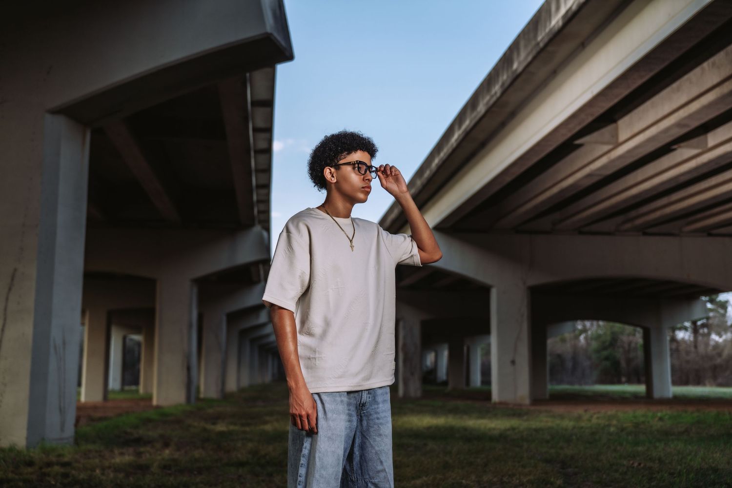 Teenage boy in white oversized shirt and jeans posing under concrete highway overpass.