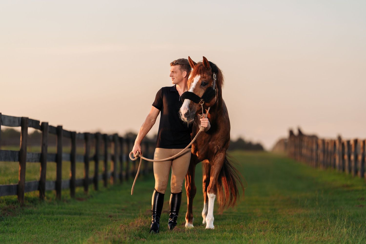 Woman in equestrian attire walking with a chestnut horse along a fenced path at golden hour.