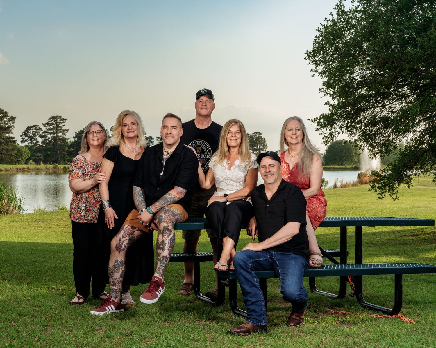 Group of seven people posing for a family portrait on a park bench near a scenic lake at sunset.