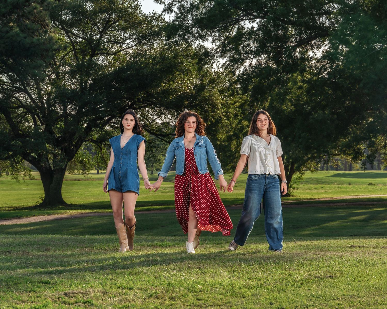 Three women holding hands walking together across a green lawn with large trees in background.