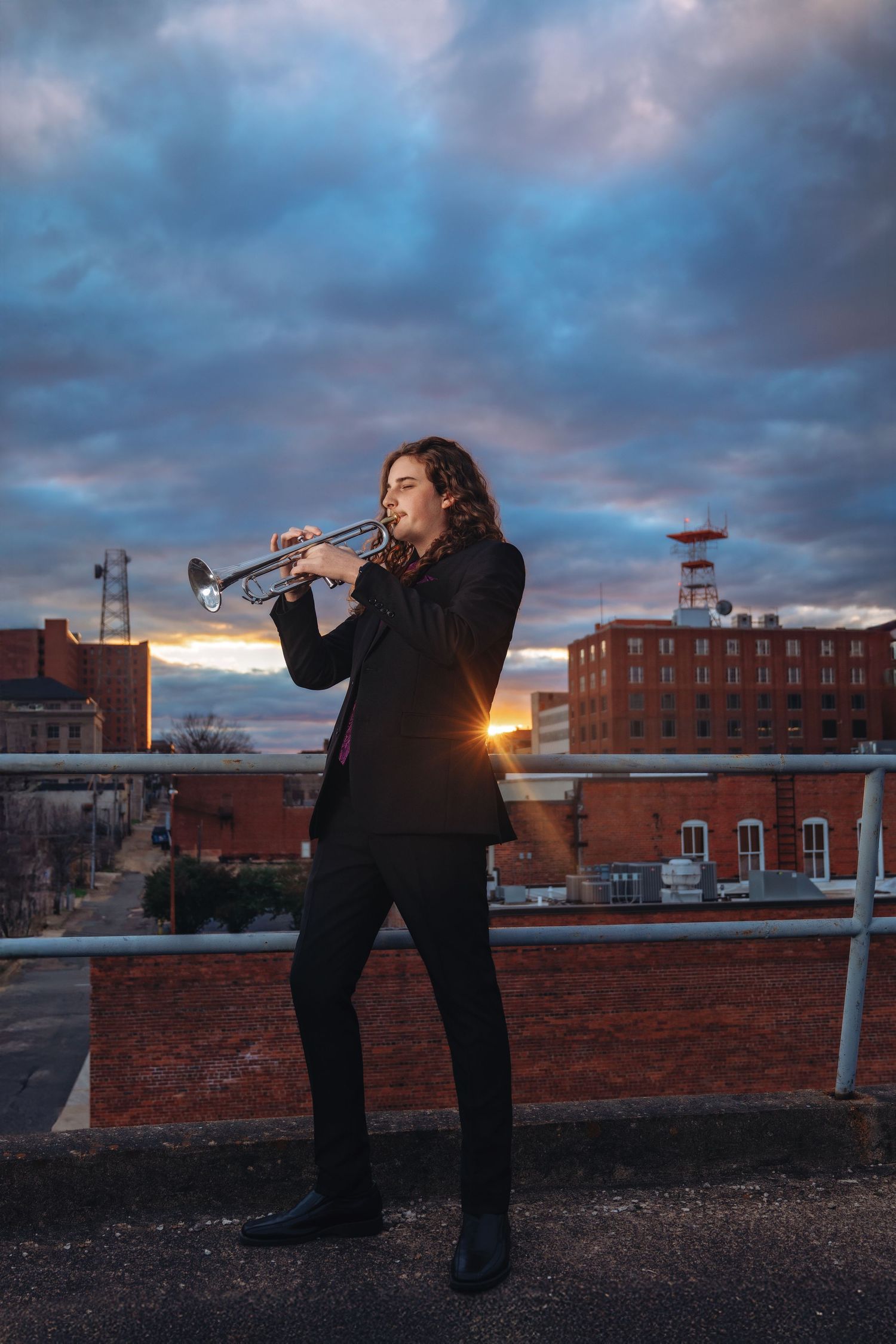 Long-haired musician in black suit plays trumpet on rooftop against dramatic cloudy sunset sky.
