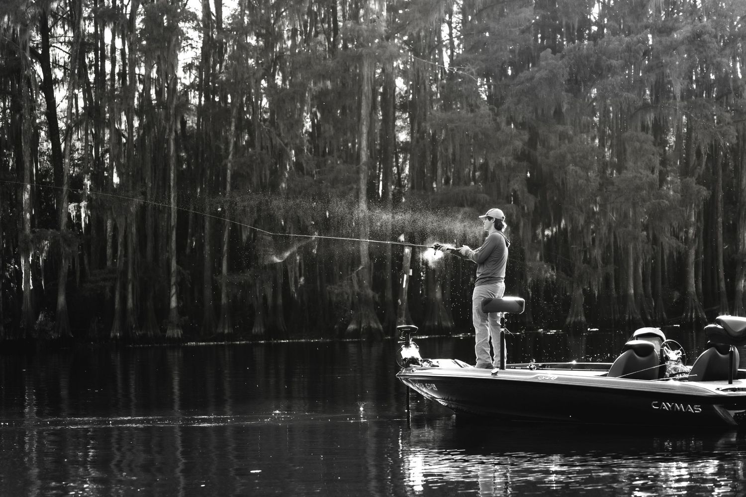 Black and white photo of a fisherman casting a line from a bass boat surrounded by cypress trees on a serene lake.