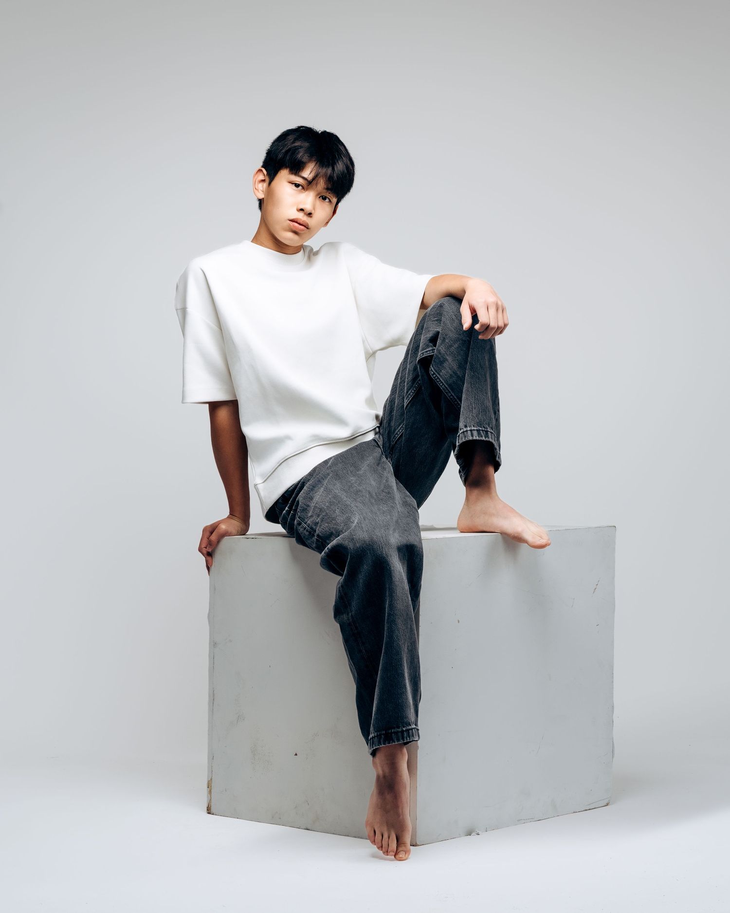 Young man in white oversized tee and dark jeans sitting barefoot on a white cube in a studio.