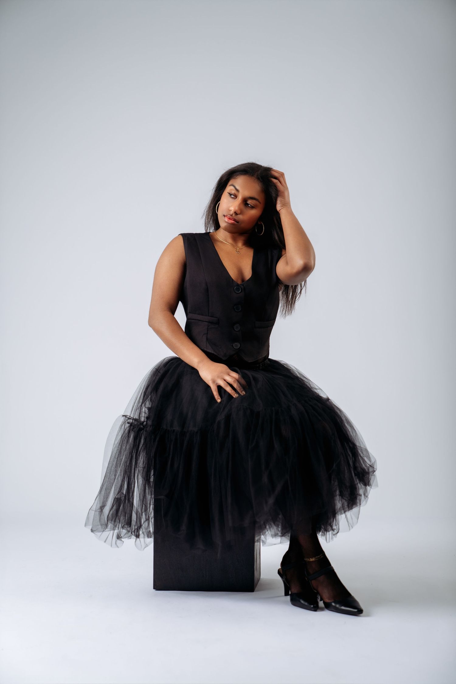 Woman in black tulle midi dress and heels sits on cube, posing with hand in hair against white background.