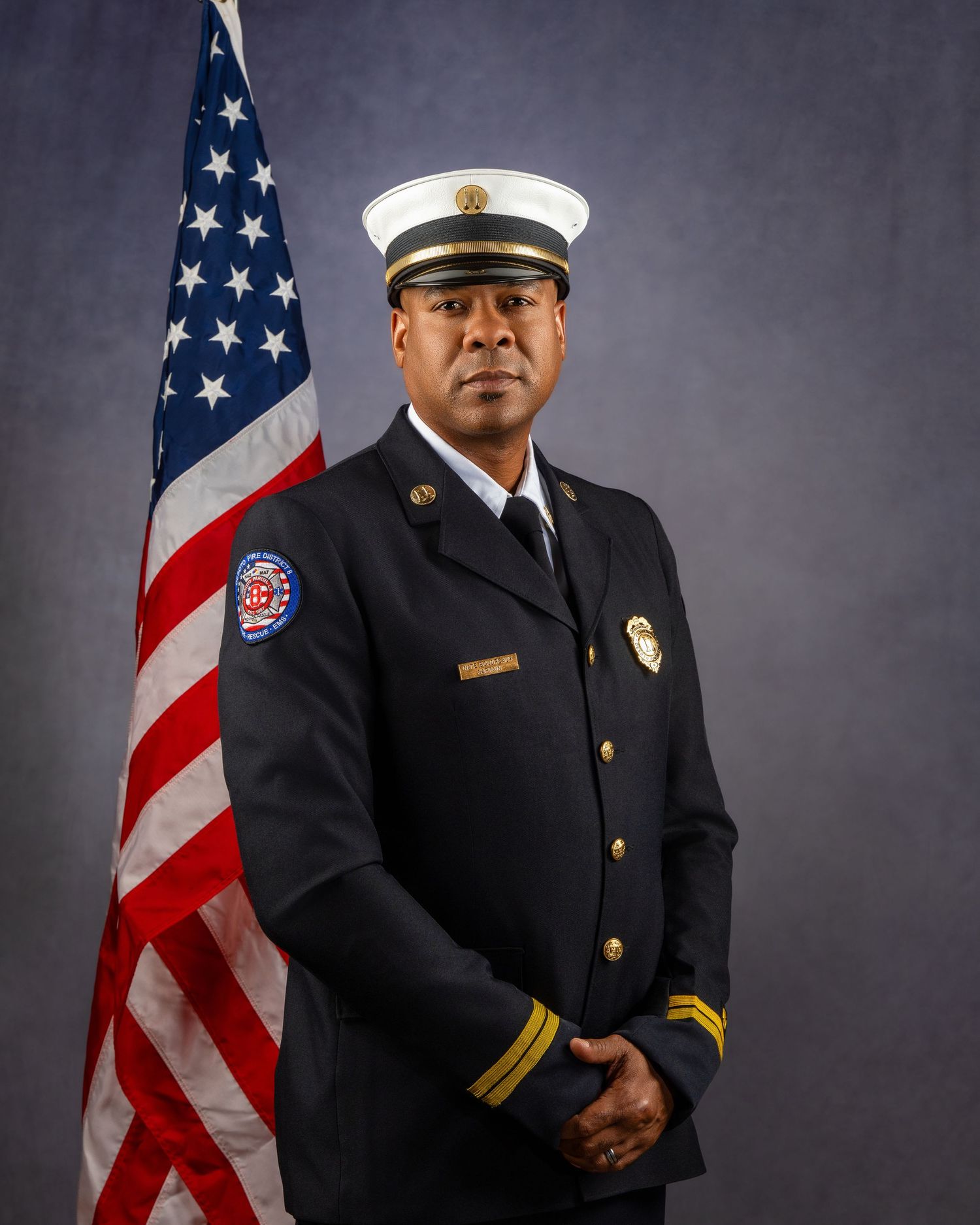 Fire department officer in dress uniform poses beside an American flag for an official portrait photo.
