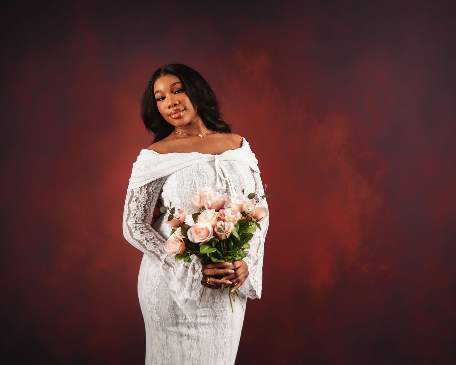 Woman in white lace off-shoulder gown holding pink roses bouquet against deep red backdrop.