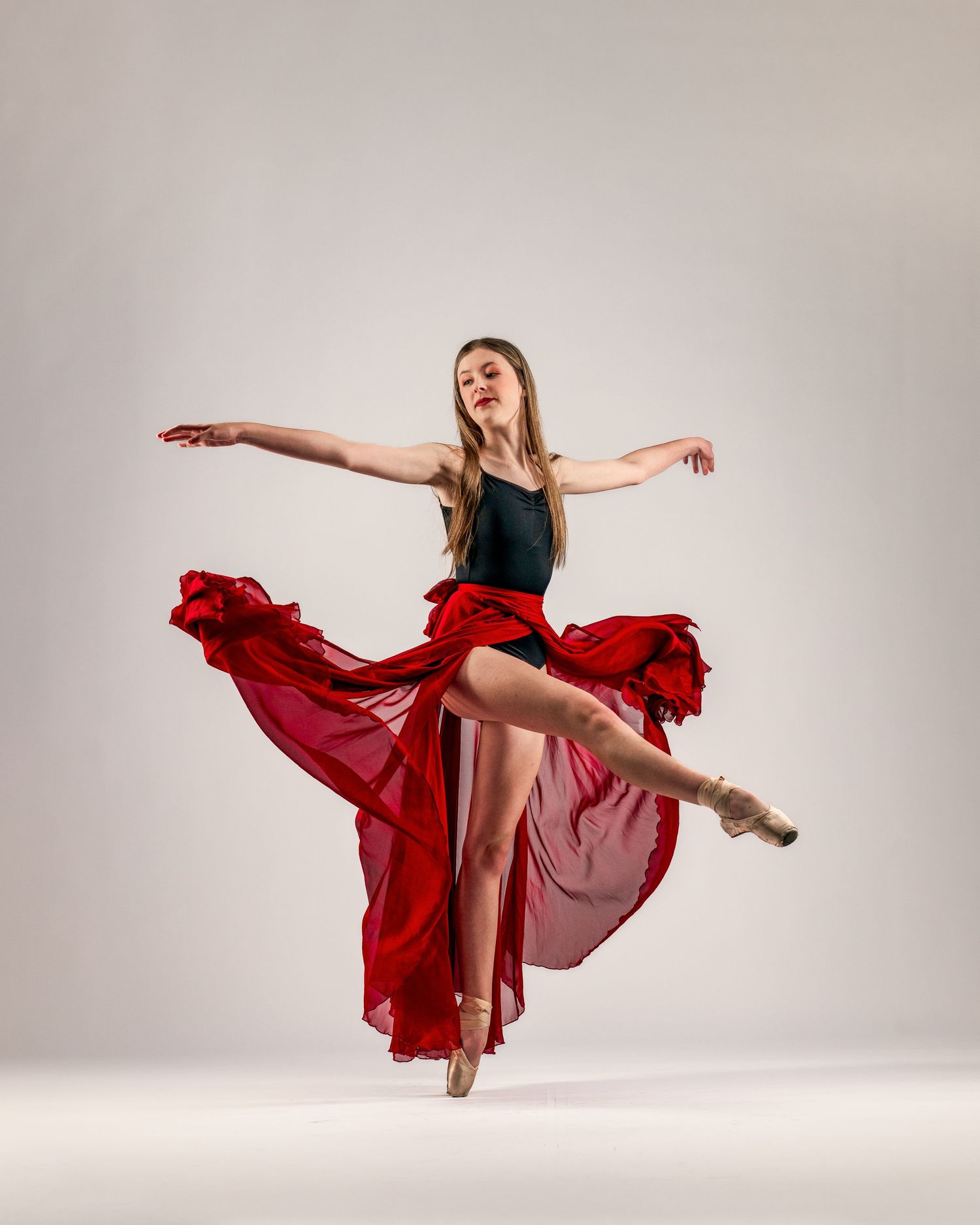 A ballet dancer in a black leotard and flowing red skirt performs an elegant pose on pointe shoes.