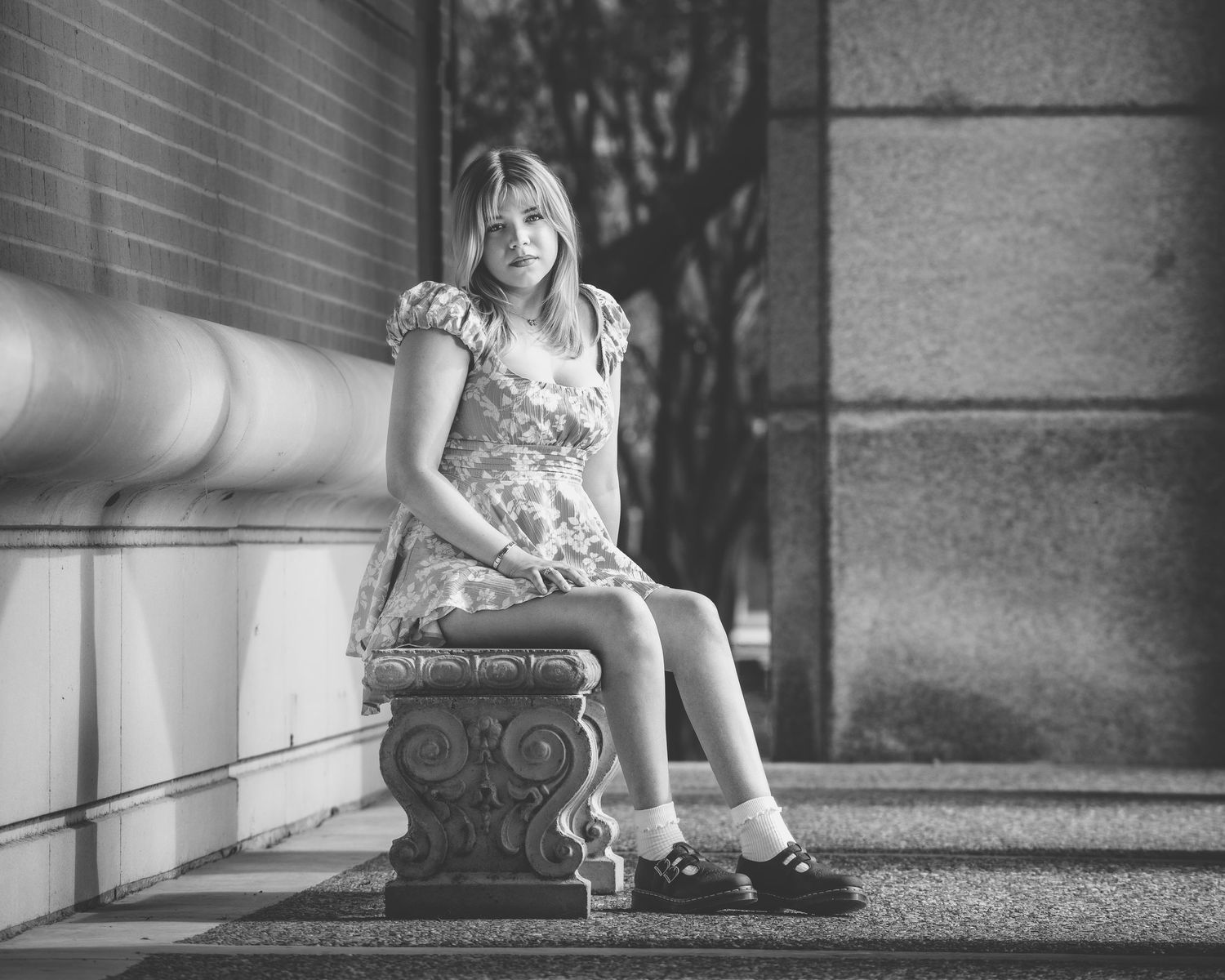 Young woman in floral dress sitting on ornate stone pedestal, black and white urban photography.