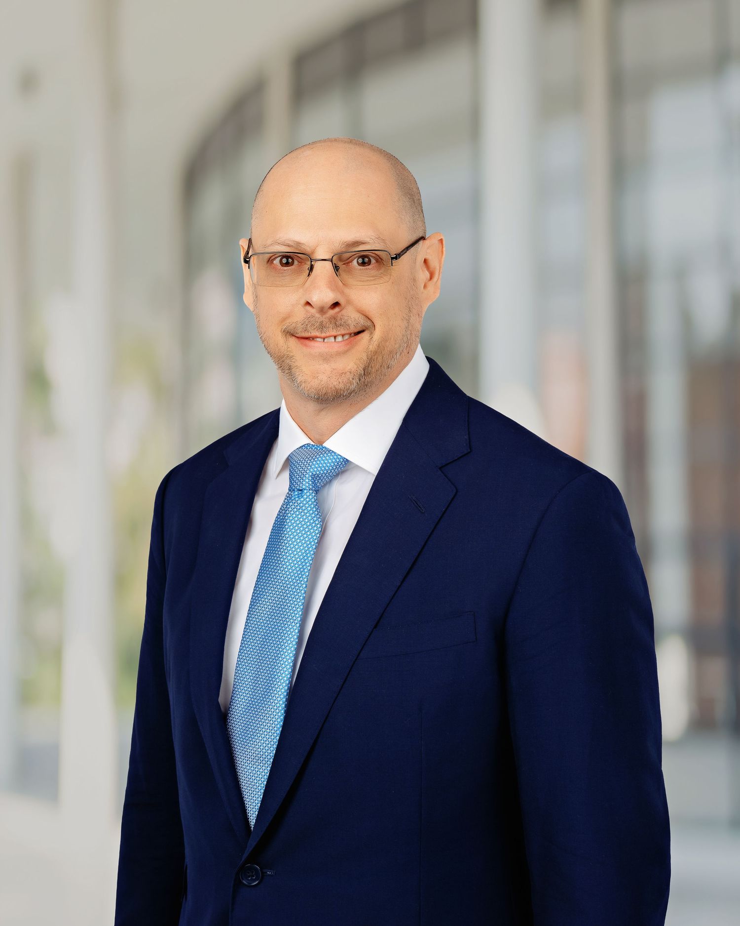 Professional bald man with glasses wearing a navy blue suit and light blue tie poses for a corporate headshot.