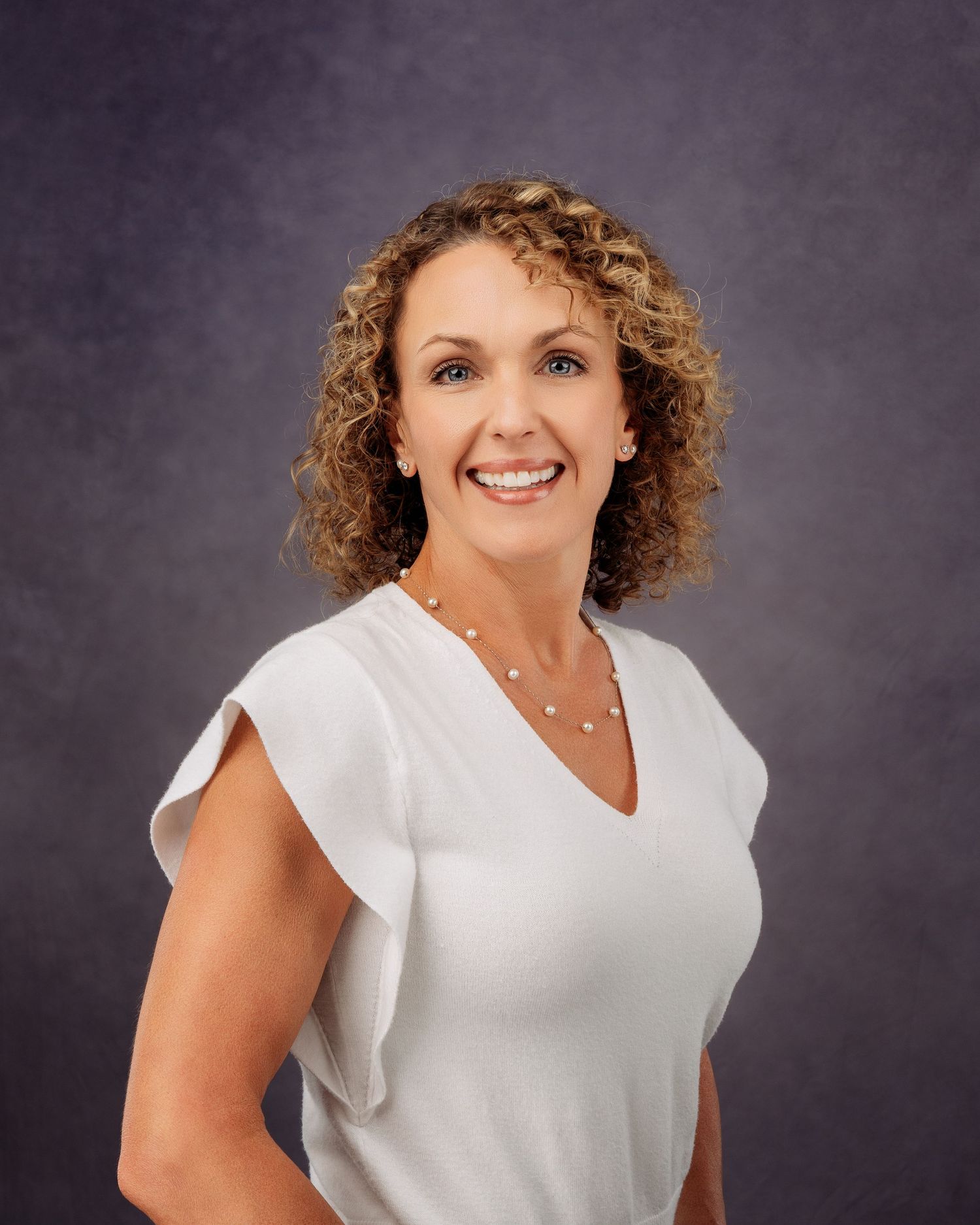 Professional headshot of a smiling woman with curly hair wearing a white top and pearl necklace against a gray background.