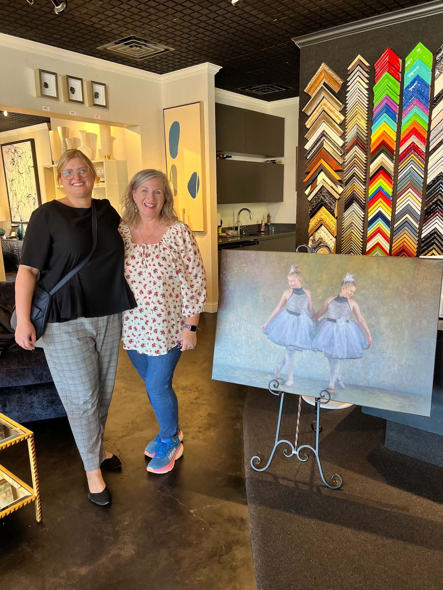 Two women stand beside a painting of two ballerinas in blue tutus on an easel in a frame shop.