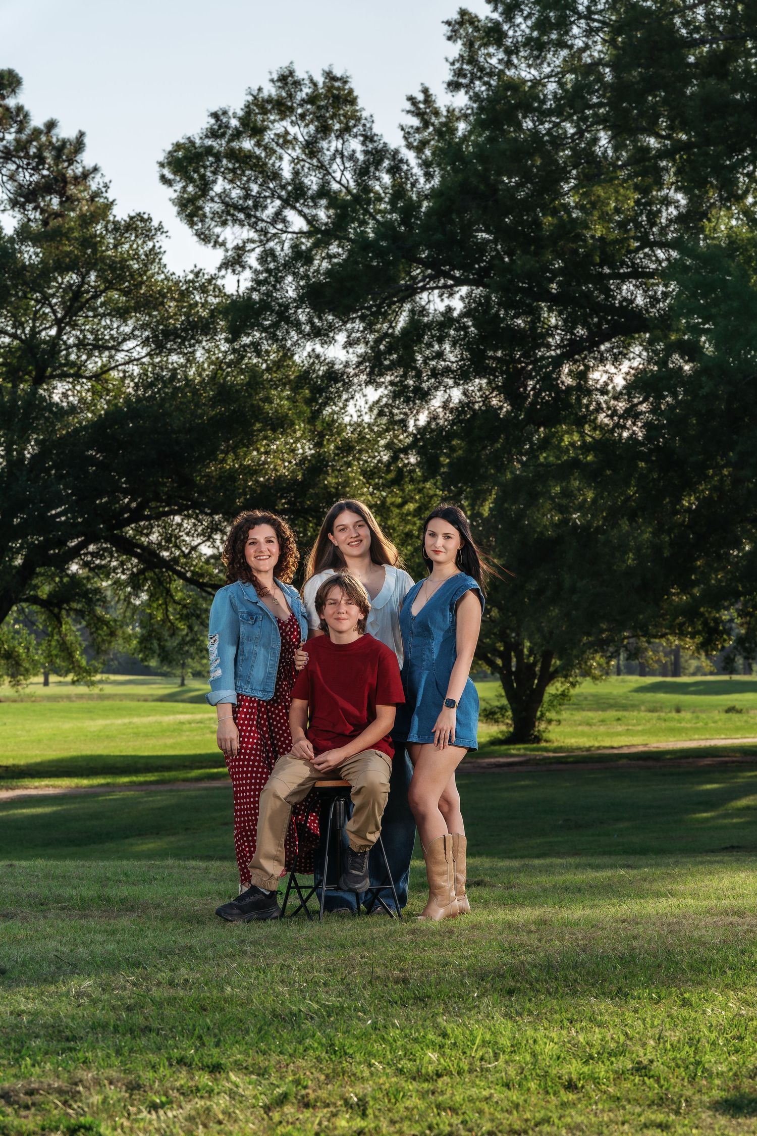 Group of four young people posing outdoors on green grass with large trees in golden sunlight.