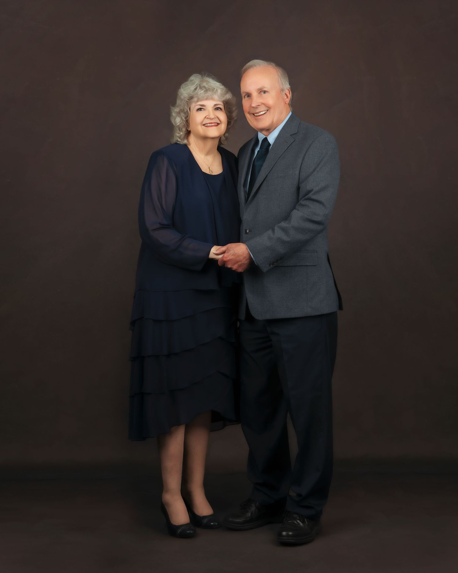Elegant older couple posing together in formal attire, woman in navy dress and man in gray suit, studio portrait.