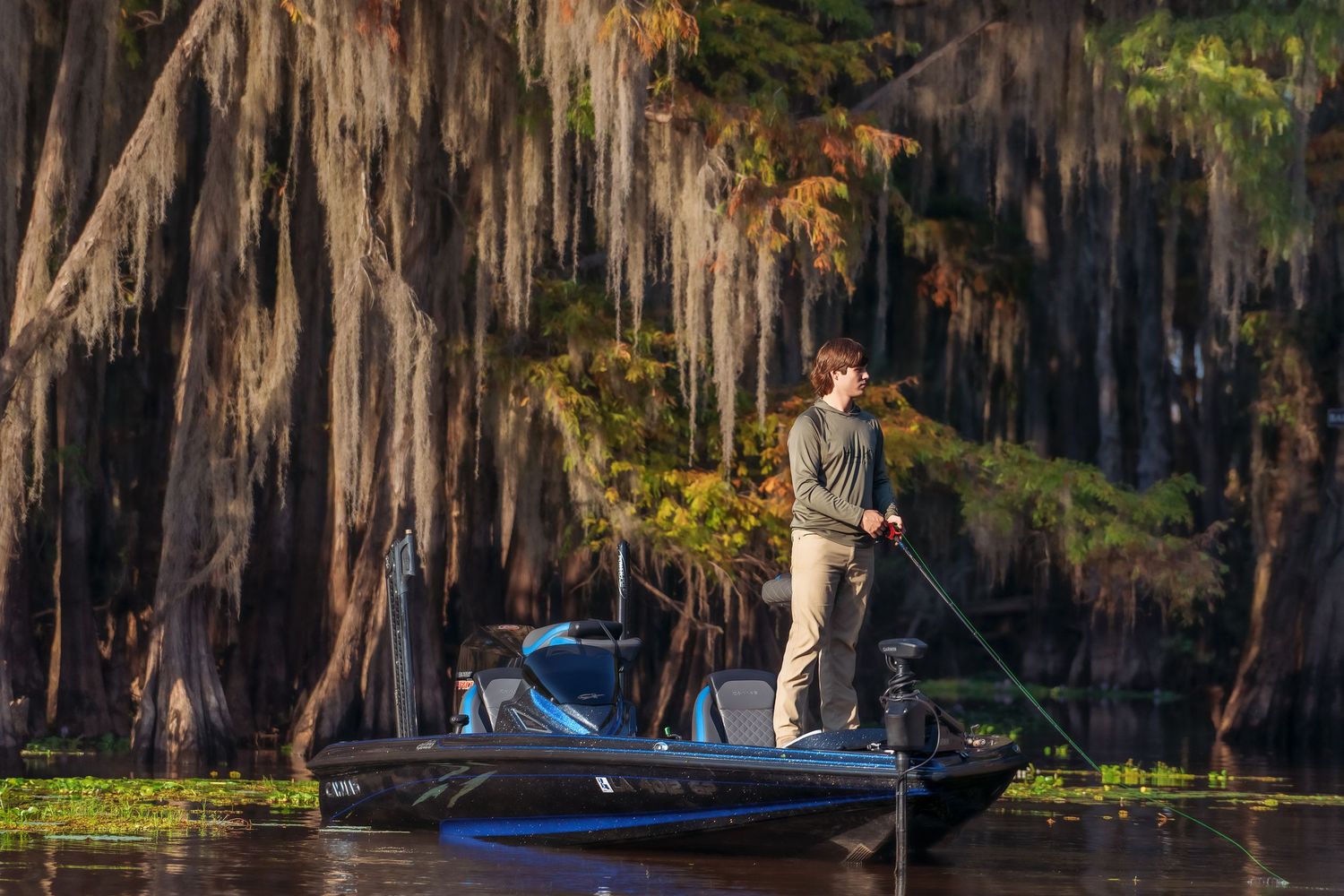 Man standing on a bass boat fishing among moss-draped cypress trees in golden light.