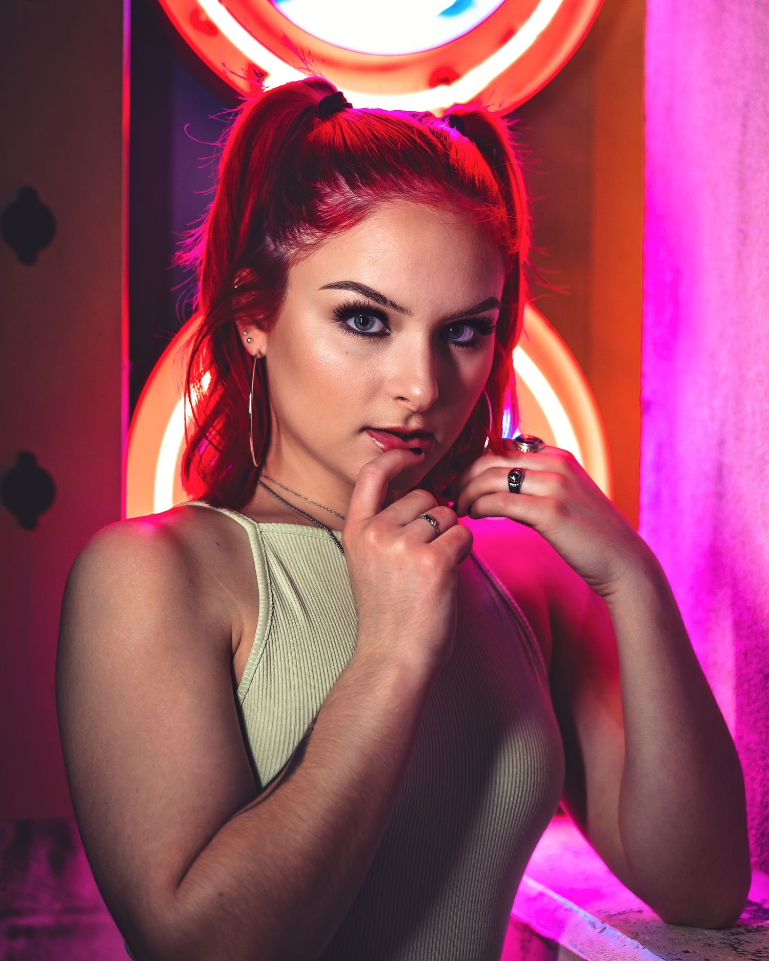 Young woman with red pigtails posing in front of neon lights in a vibrant pink-lit setting.