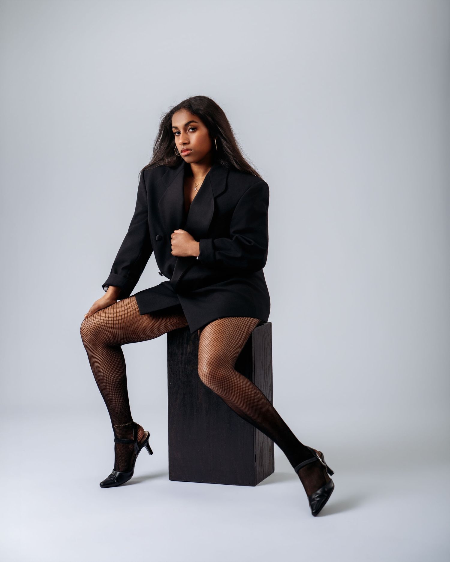 A young woman in an all-black ensemble with fishnet stockings sits on a dark cube in a studio setting.