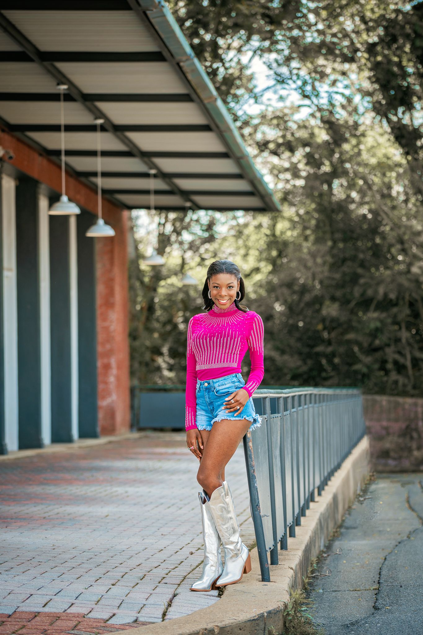 Young woman in pink top, denim shorts, and white cowboy boots poses smiling on a covered walkway with railing.