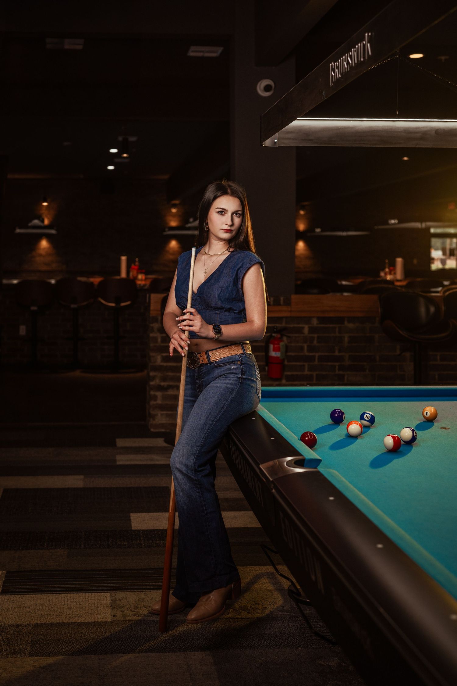 Confident woman holding a billiard cue leaning against a pool table in a bar.