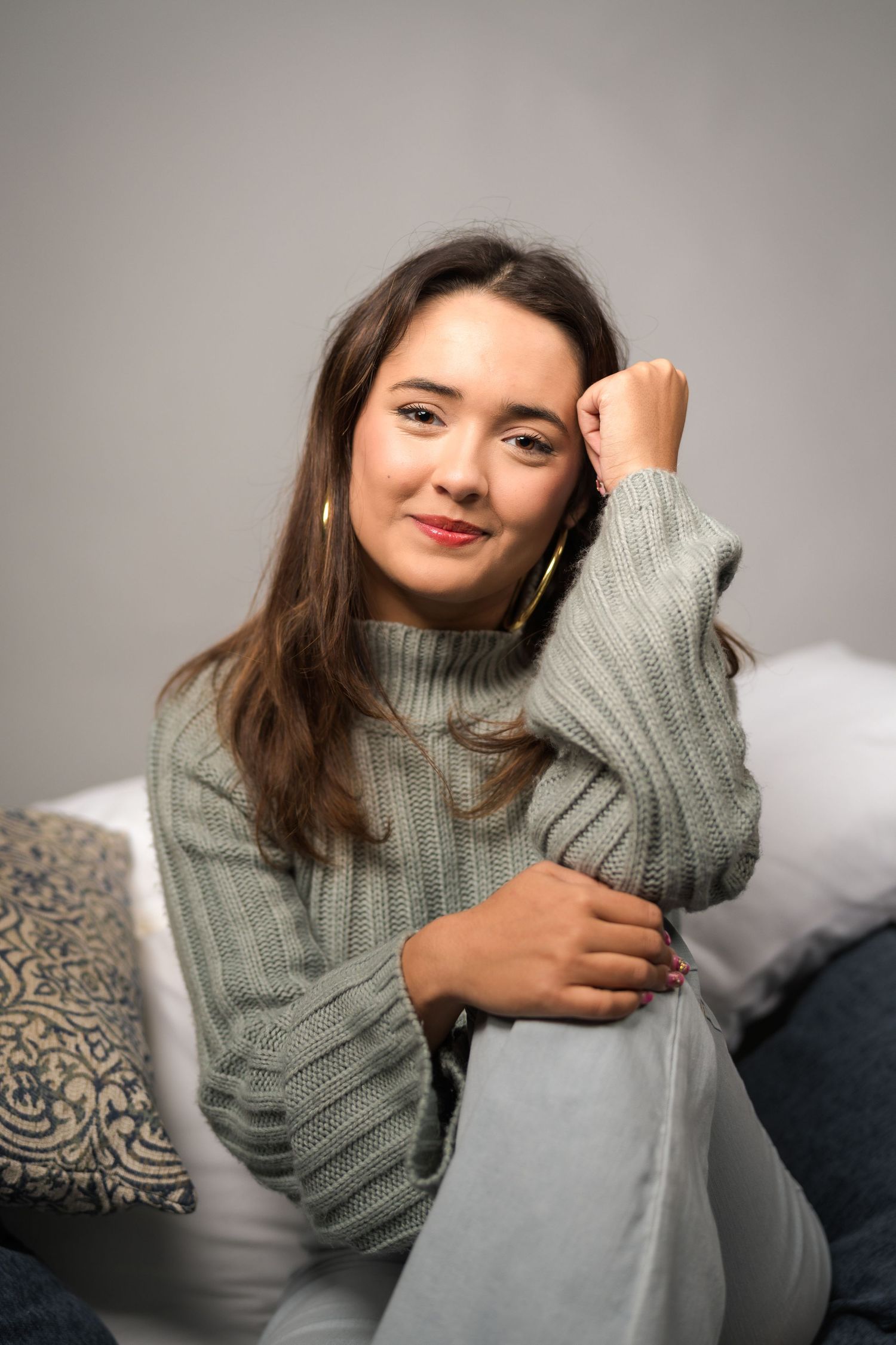 Young woman in sage green knit sweater sitting relaxed on couch with decorative pillows, smiling softly.