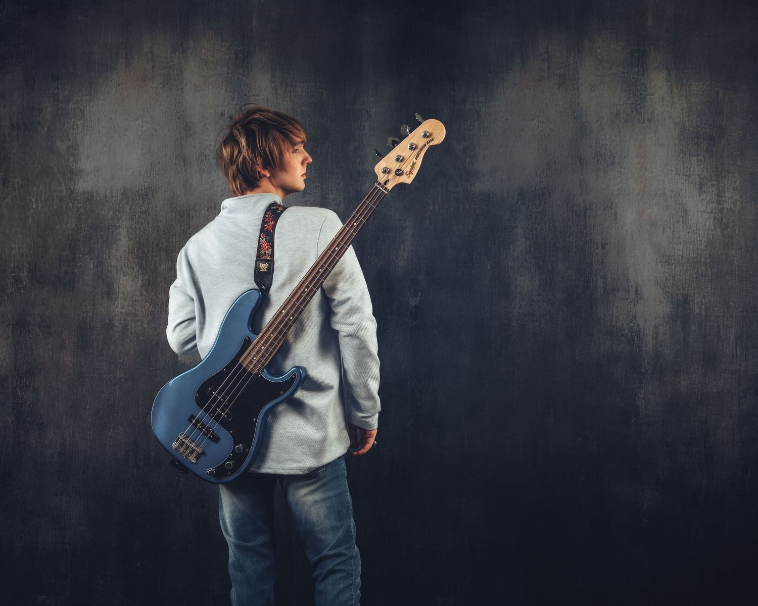 A young male guitarist stands with his back turned, holding a blue electric bass guitar against a dark gray background.