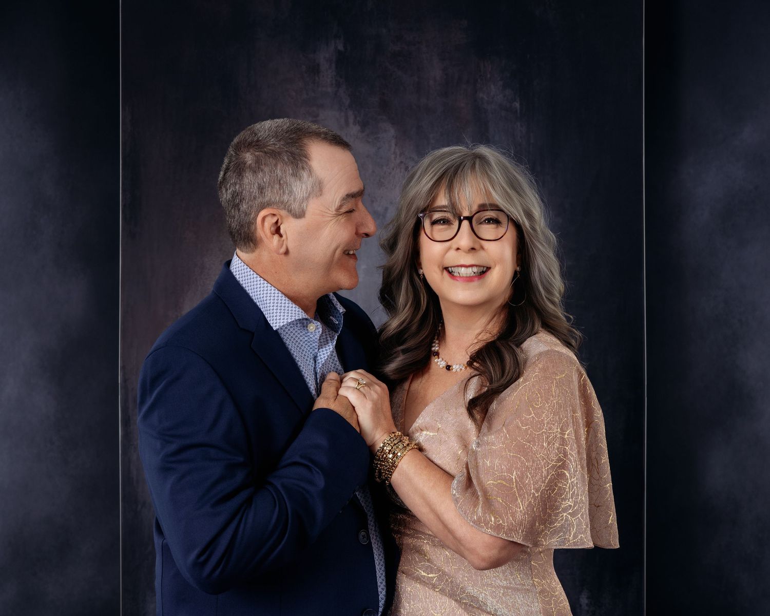 A smiling couple poses together, the man in a navy blazer and woman in a champagne dress with glasses, against a dark backdrop.