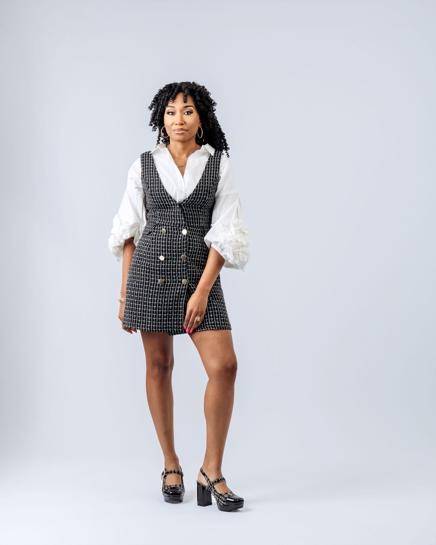 Woman wearing a black tweed double-breasted mini dress over a white puff-sleeve blouse with black platform heels.