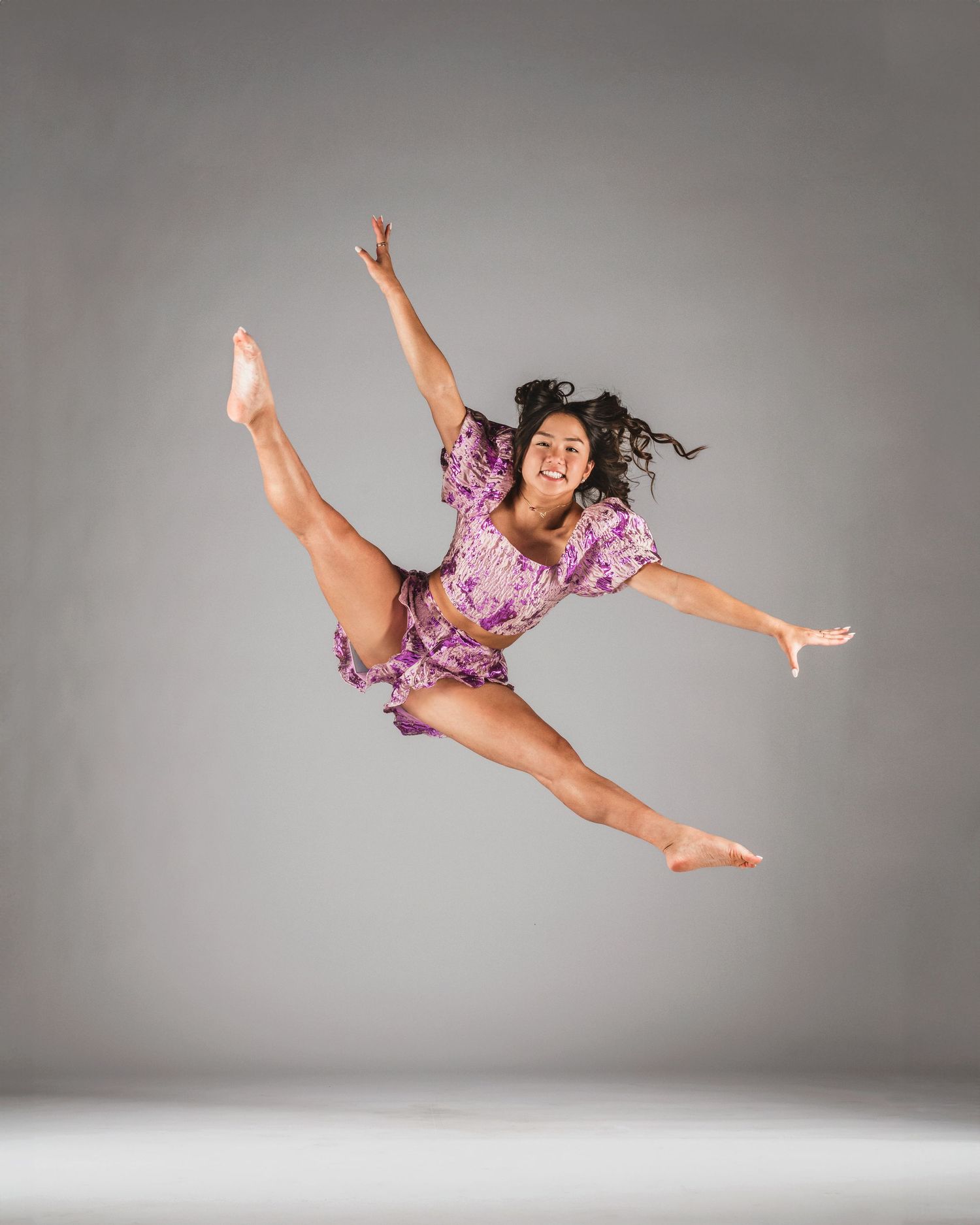Dancer in purple floral outfit leaping mid-air with legs split and arms extended against gray background.