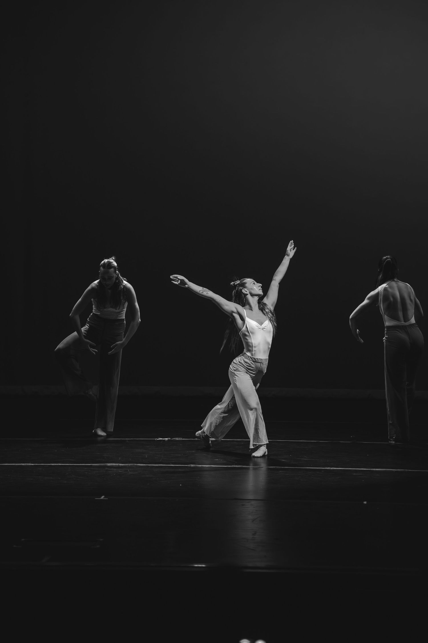 Three dancers perform on a dimly lit stage in black and white, with one striking a graceful pose with arms raised.
