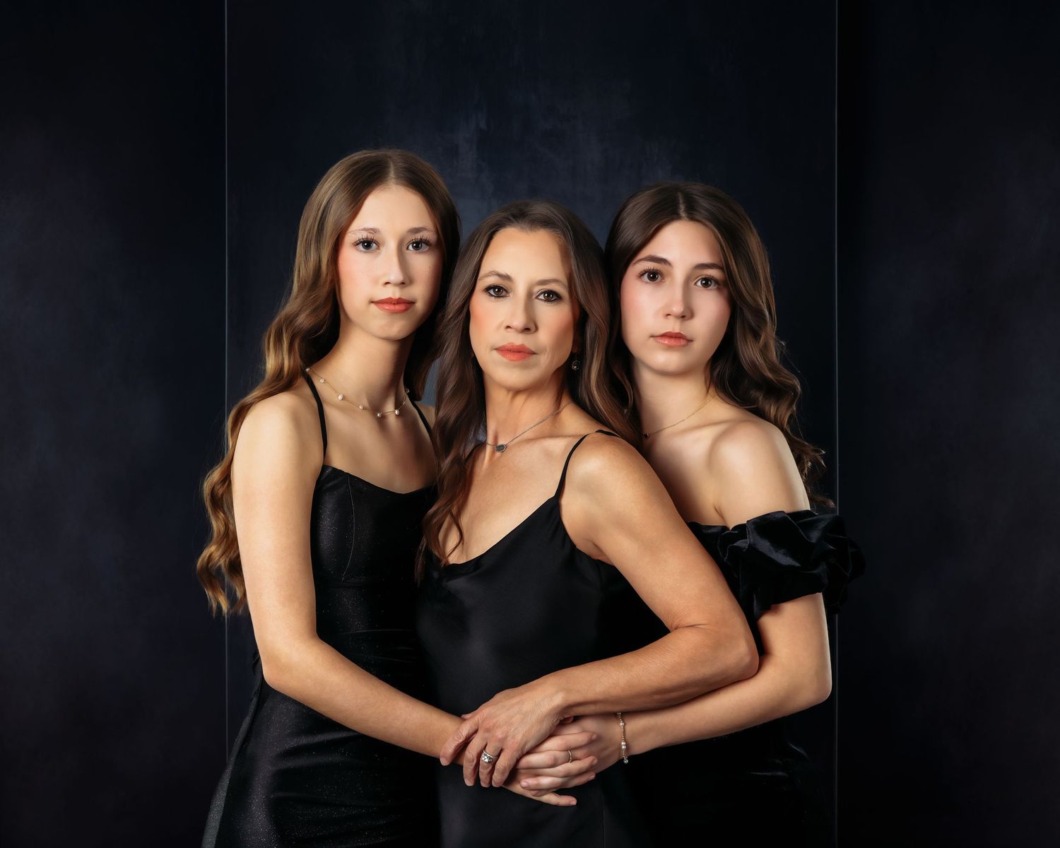 Three women in elegant black dresses pose together for a formal portrait against a dark blue backdrop.