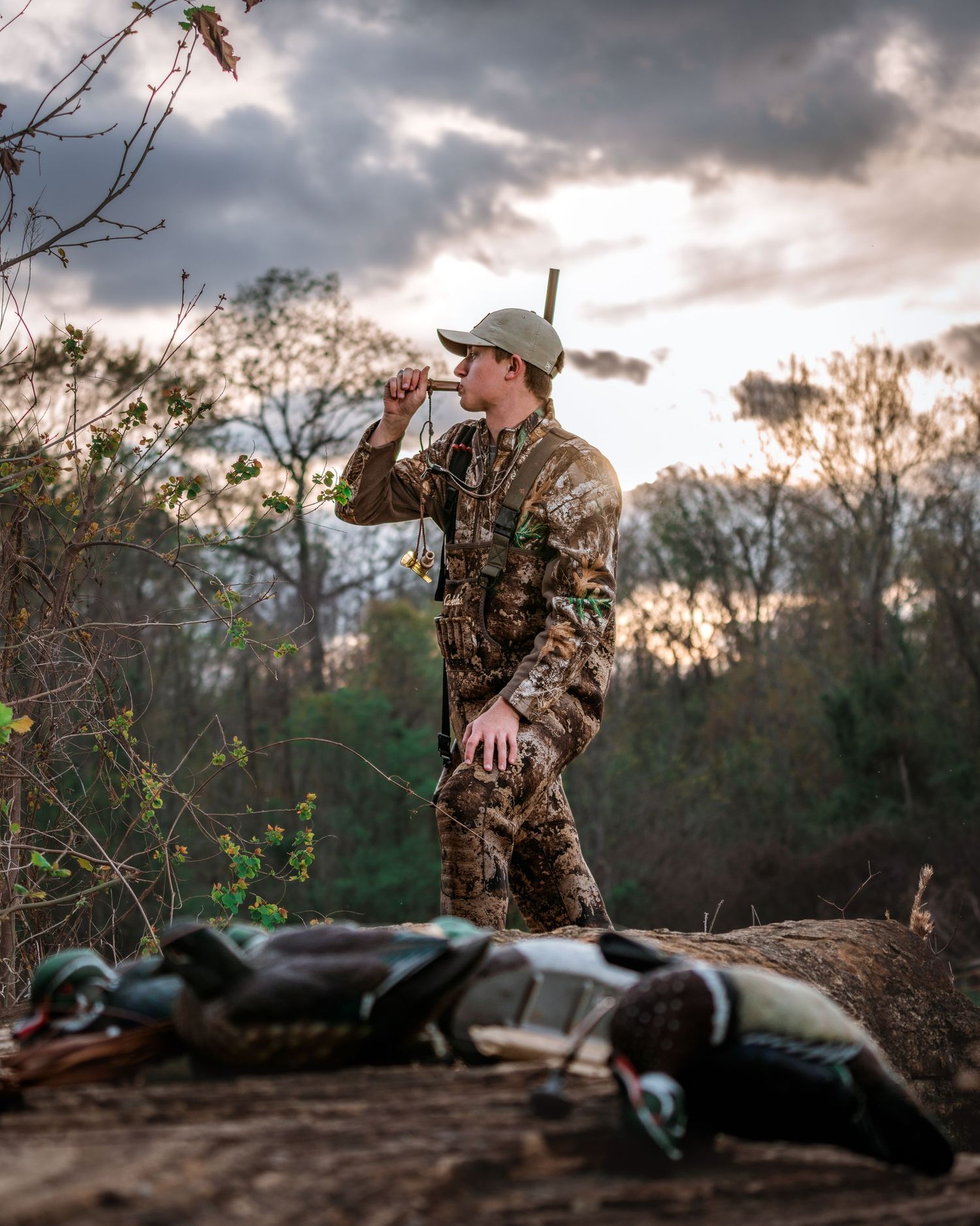 Hunter in camouflage gear blowing a duck call at dusk with decoys on the ground nearby.