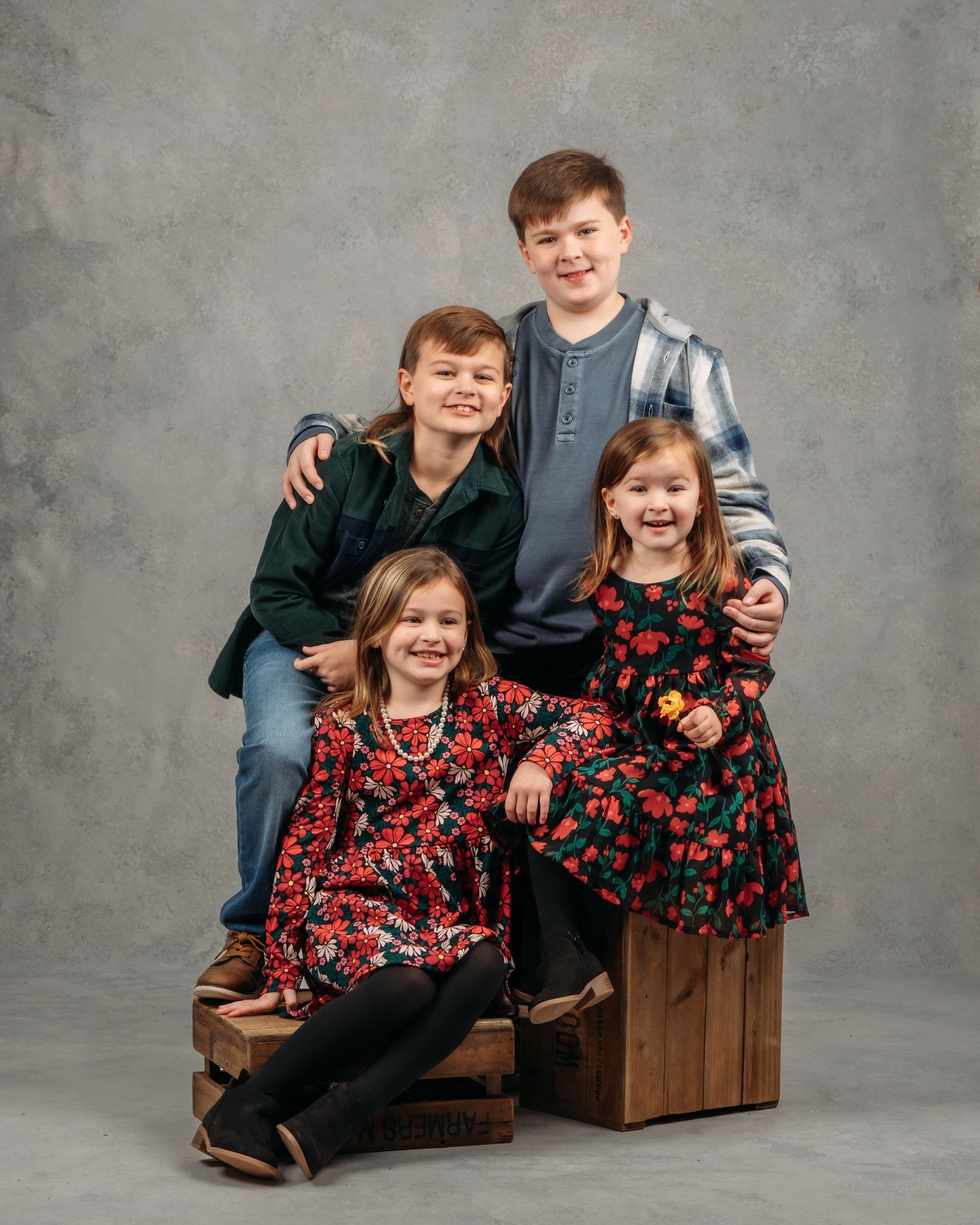 Four children smile together in a studio portrait, seated and standing on wooden crates.