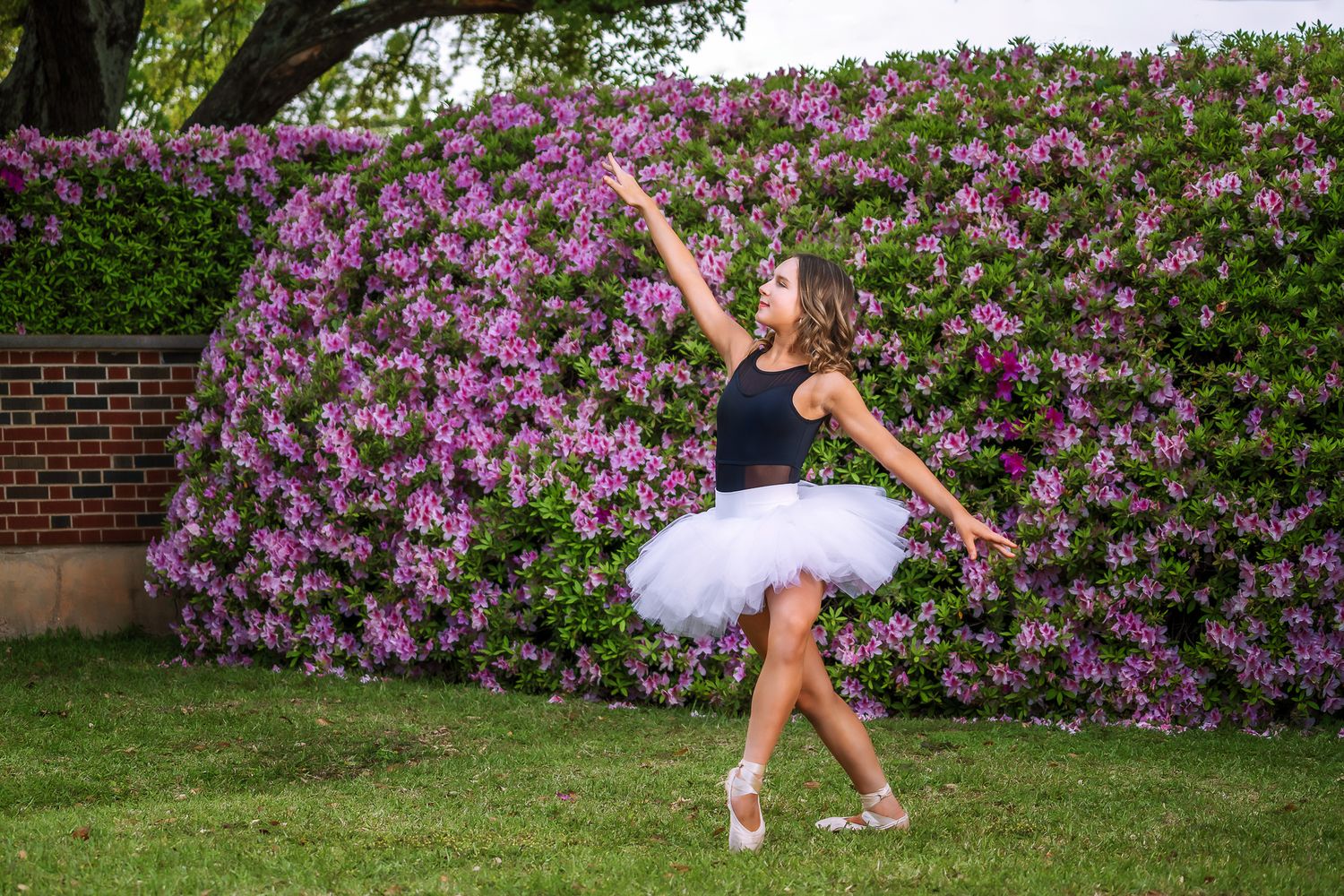 Ballerina in white tutu performing arabesque outdoors in front of blooming purple flower wall.