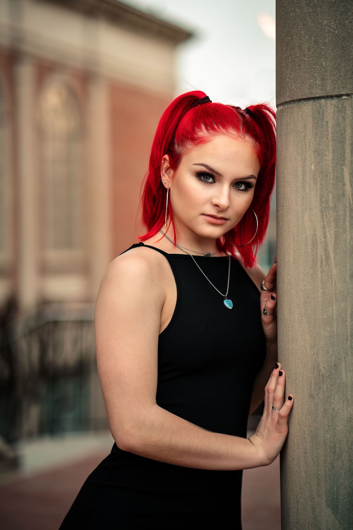 Young woman with vibrant red pigtails in a black dress leans against a stone column outdoors.