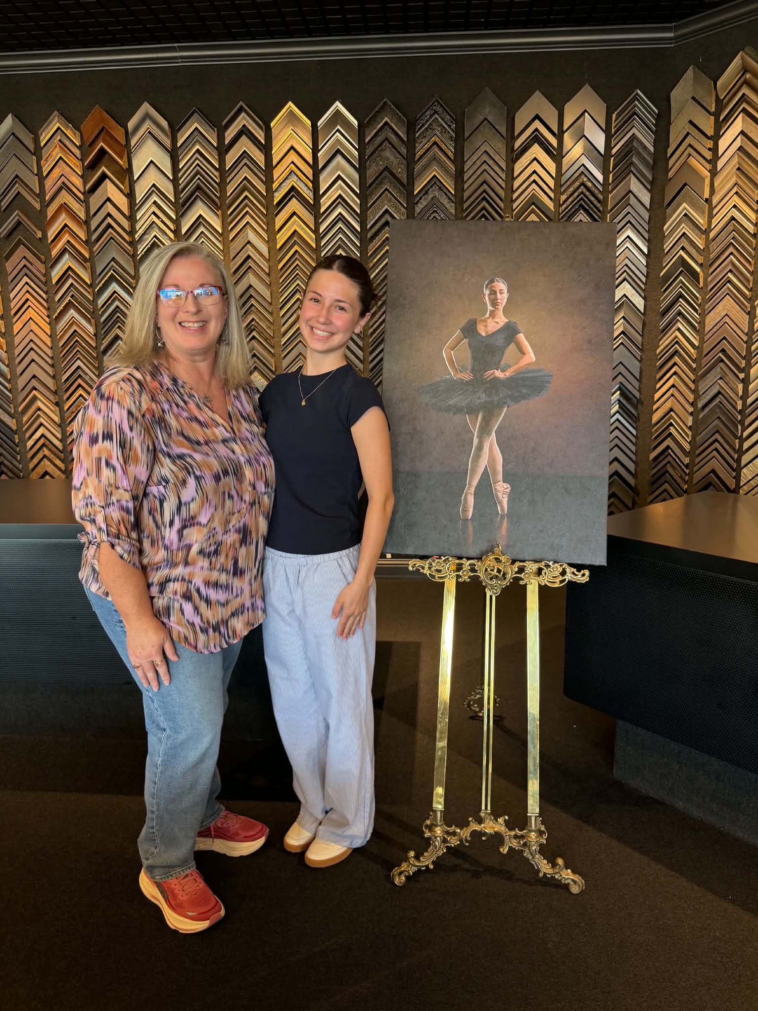 Two women smile beside a framed ballerina portrait on a gold easel in a frame shop.