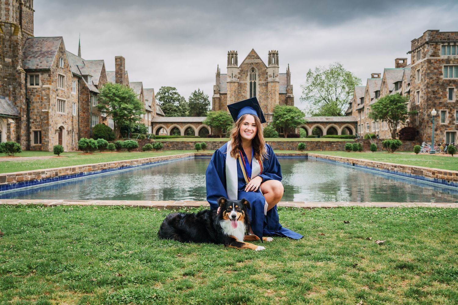 Female graduate in blue cap and gown kneeling with her dog beside a reflecting pool on campus.