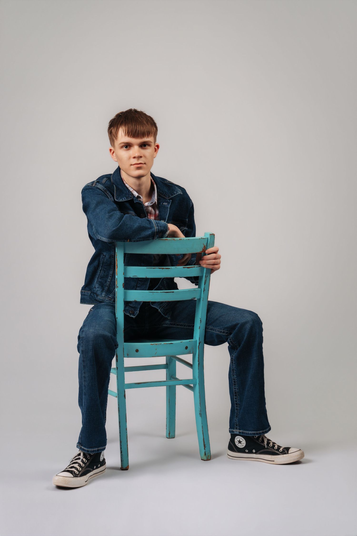 Young man in denim jacket and jeans sitting backwards on a teal wooden chair against a gray background.