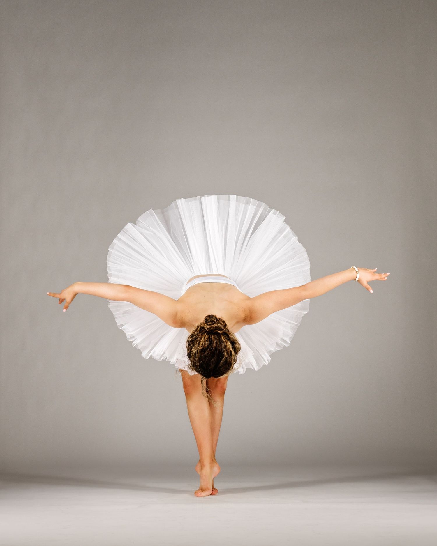 A ballerina in a white tutu bends forward gracefully with arms outstretched in a studio setting.