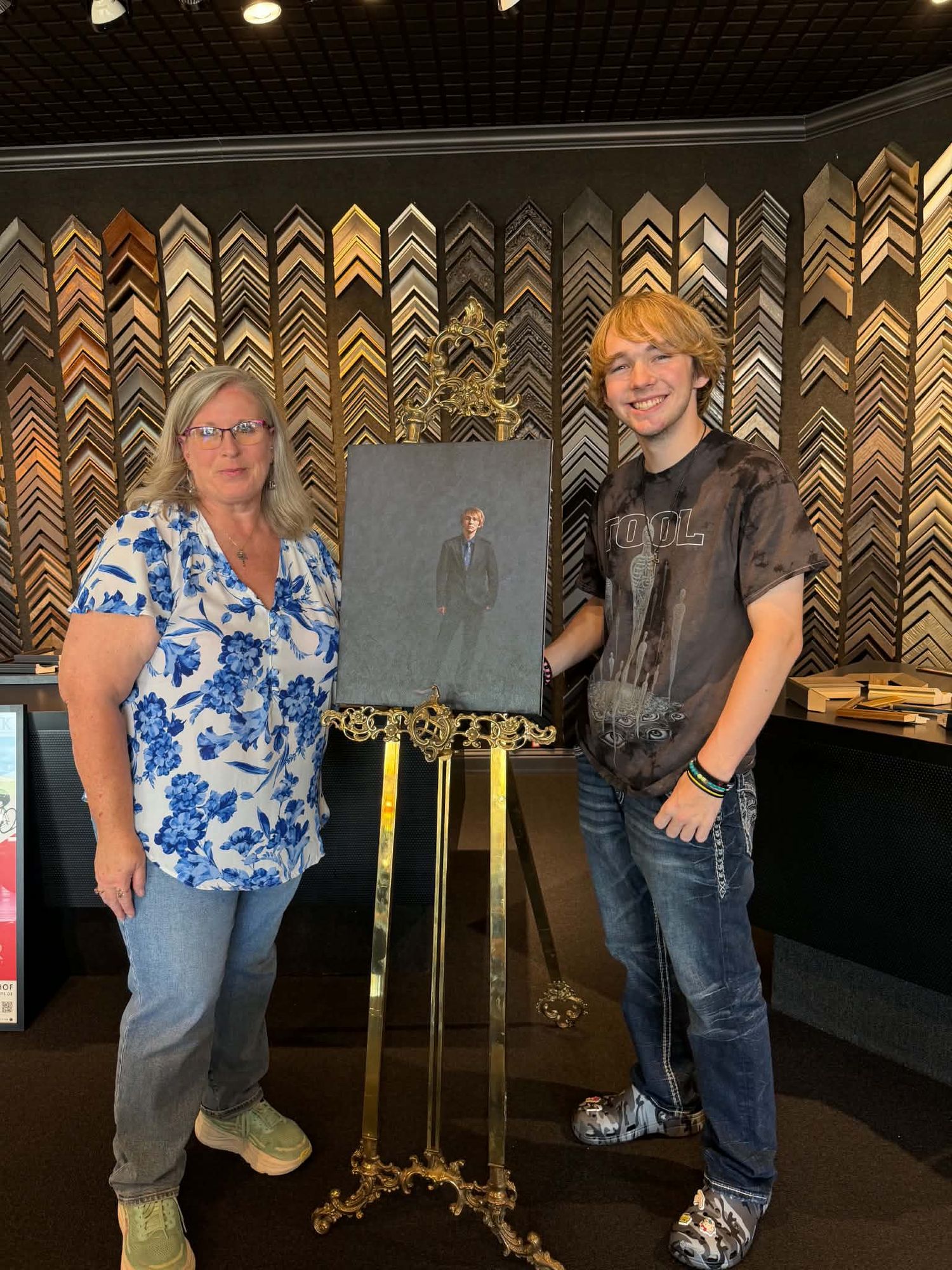 Mother and teenage son standing beside a framed portrait on an easel in a frame shop.