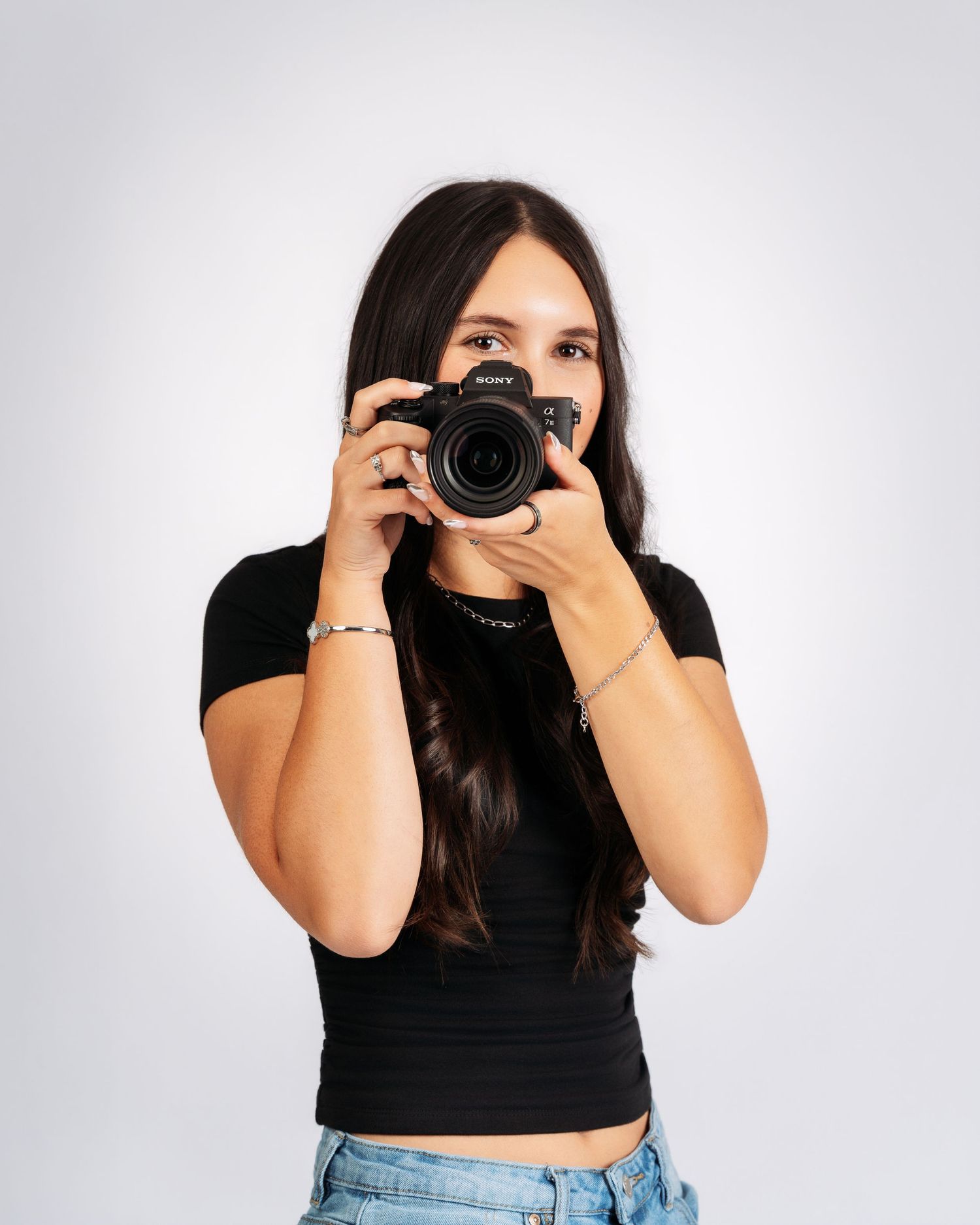 Young woman in black crop top holds Sony camera up to her face, photographing against a white background.