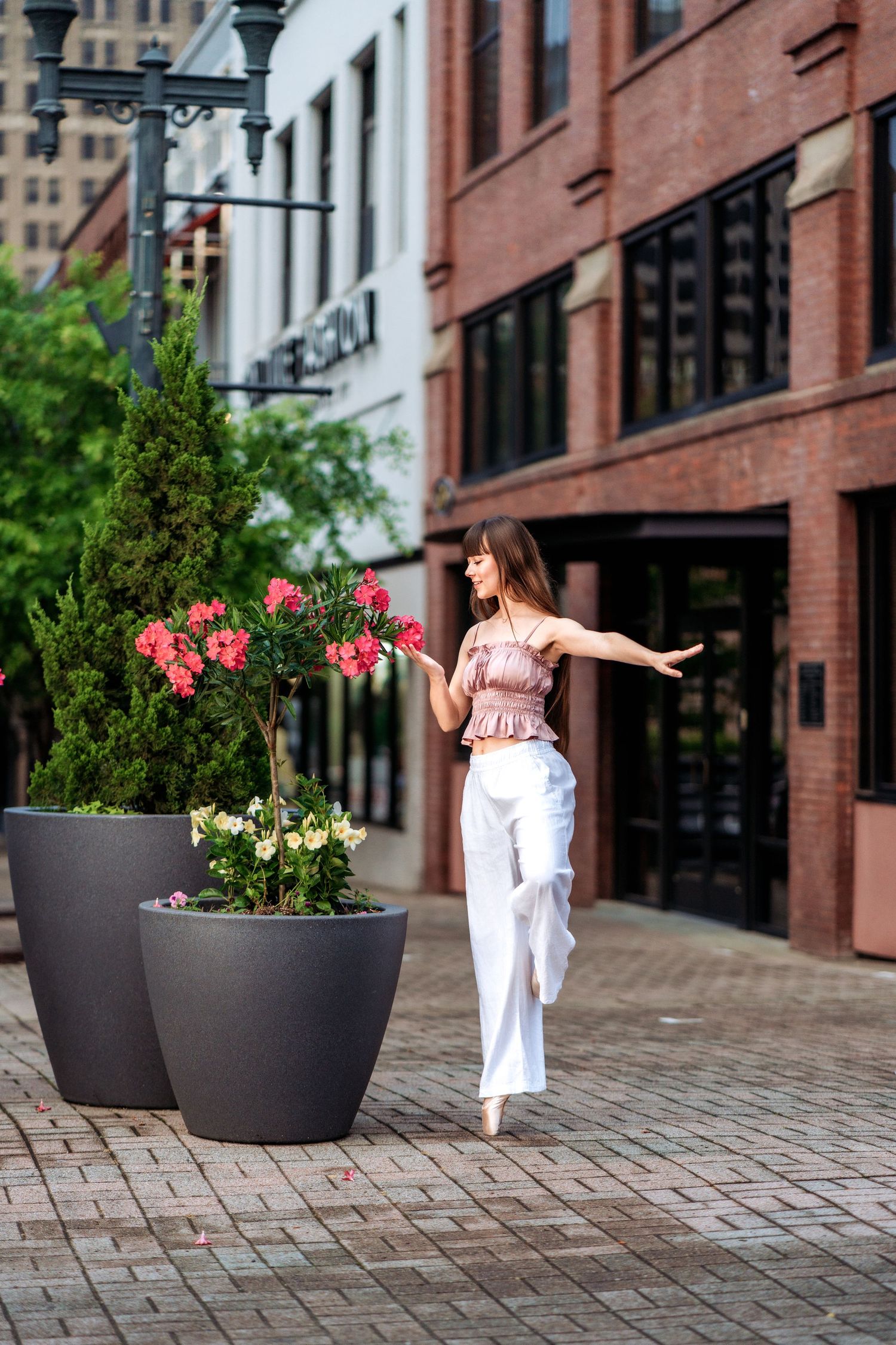 Young woman in white pants and pink top dancing gracefully on a brick sidewalk near large planters with flowers.
