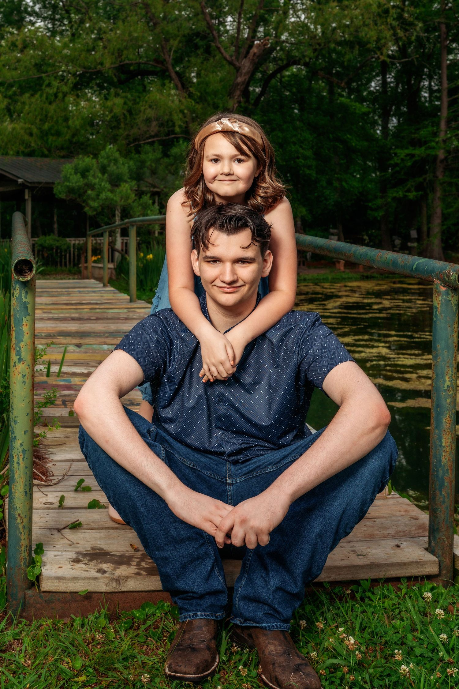 A young man sits on a dock with a smiling girl on his shoulders near a pond.