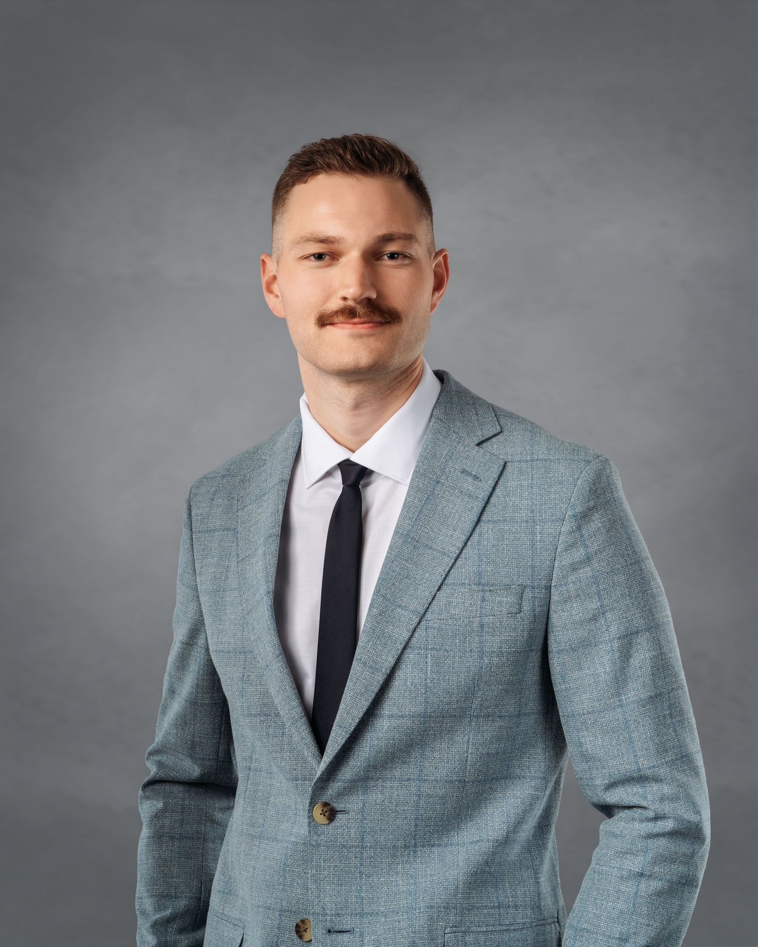 Professional man with mustache wearing light blue suit and black tie poses for formal business portrait.