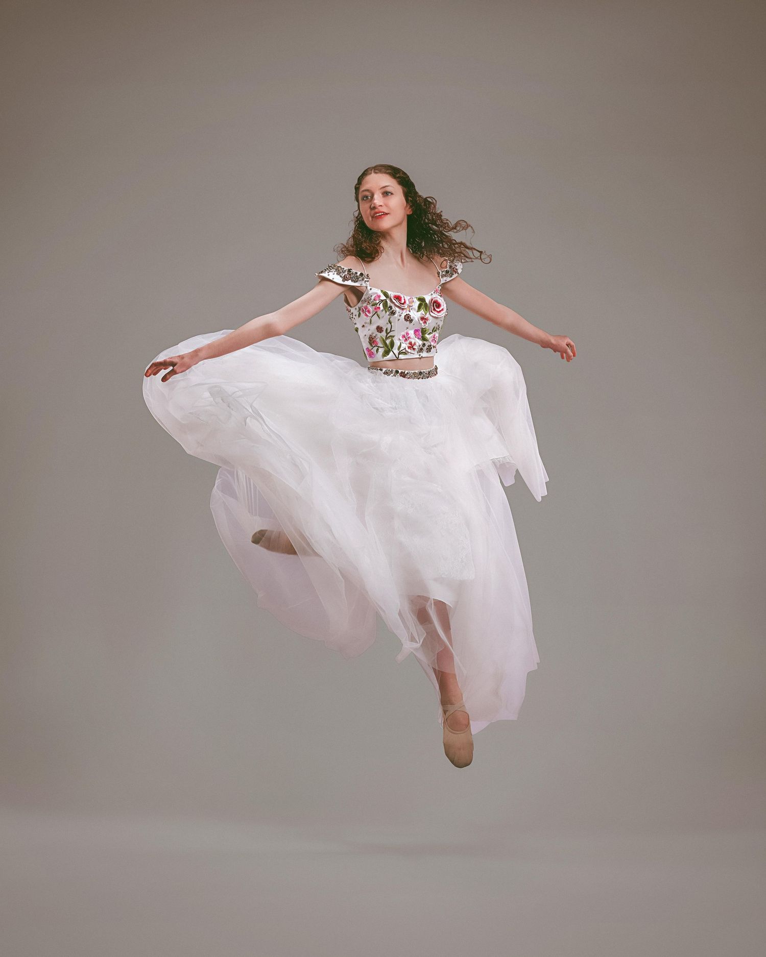 Dancer leaping gracefully in a white tulle skirt and floral embroidered crop top against a gray background.