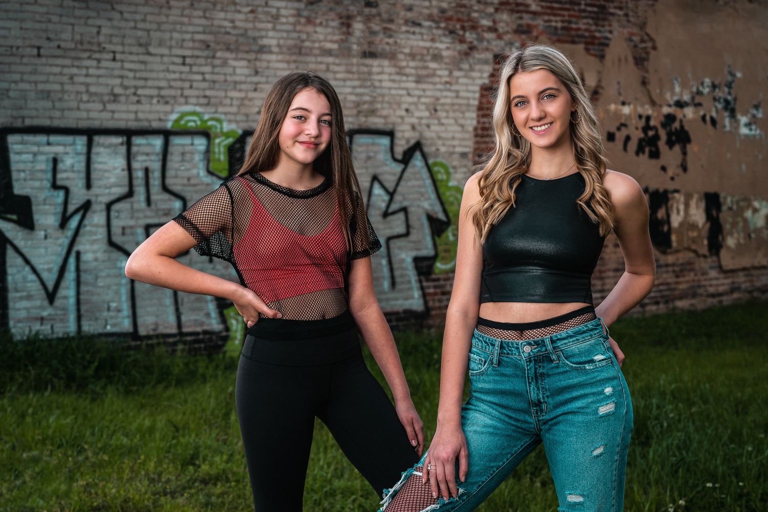 Two teenage female dancers pose together in front of graffiti wall in casual dance attire.