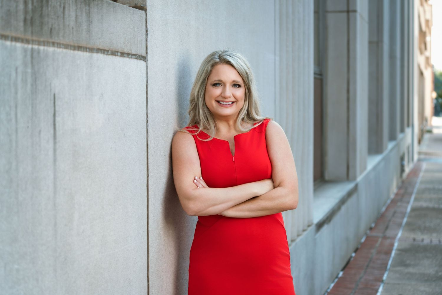 Professional woman in red dress smiling with arms crossed, leaning against a concrete wall outdoors.
