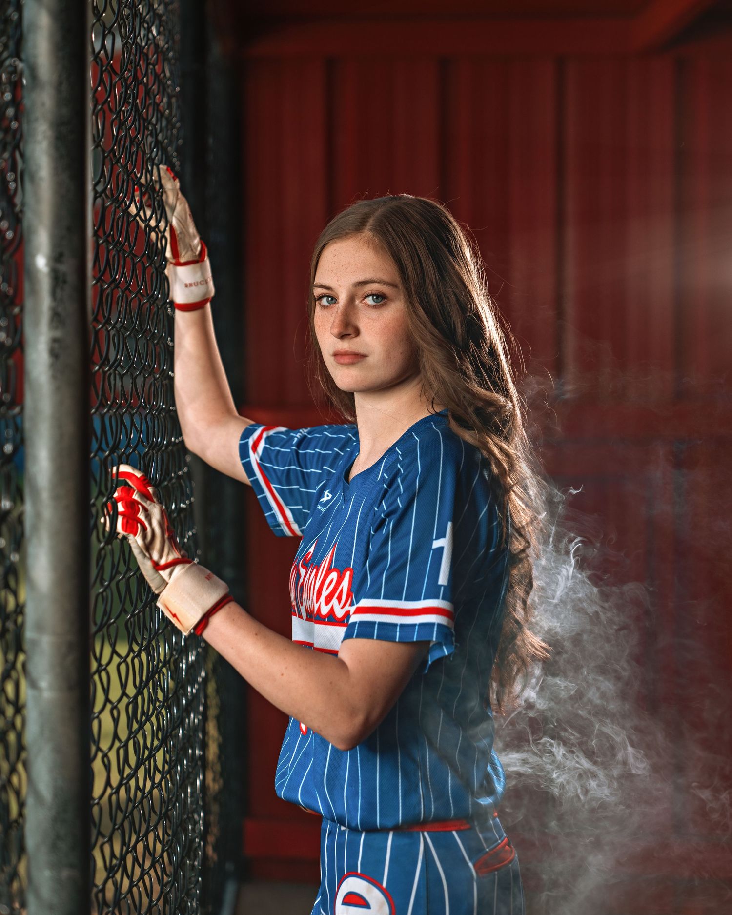 Young female softball player in blue pinstripe jersey poses confidently by chain-link fence with dramatic smoke effect.