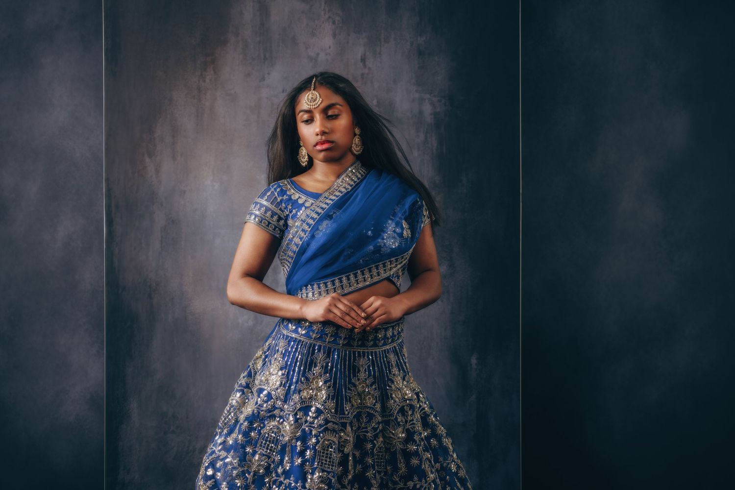 Woman in elegant blue and gold embroidered lehenga with traditional jewelry poses gracefully against dark background.