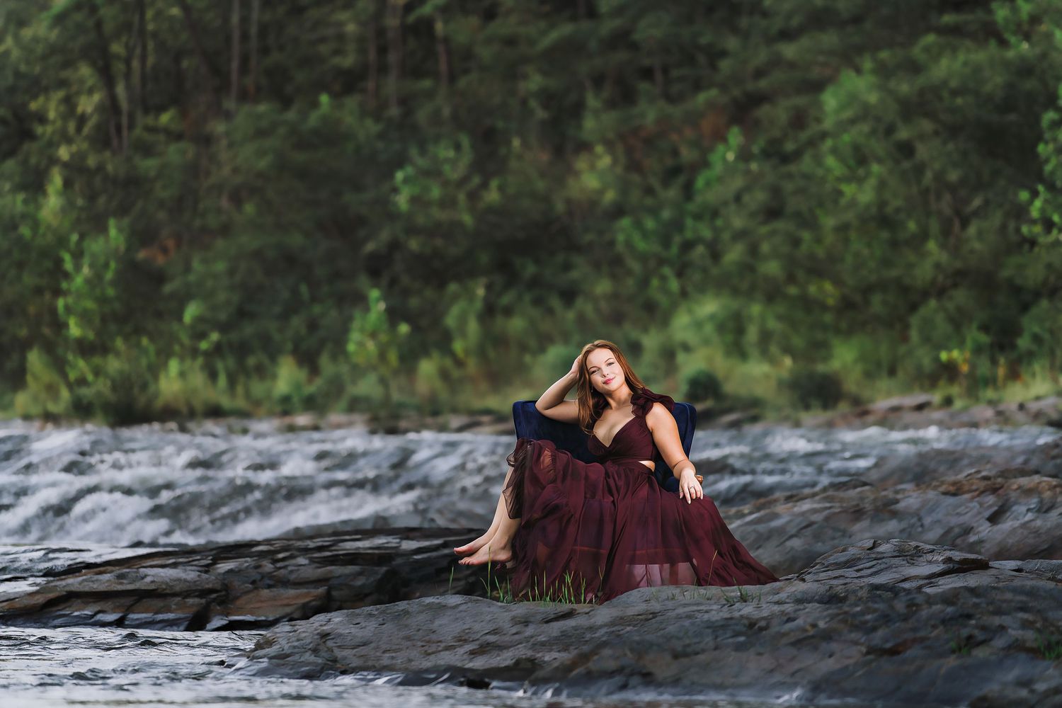 Woman in burgundy gown poses gracefully on rocky riverbank with lush green forest backdrop.